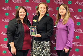  three women stand together with the woman in the middle holding an award